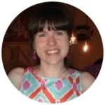 A woman with straight brown hair and bangs, wearing a colorful geometric-patterned sleeveless top, smiles at the camera indoors.