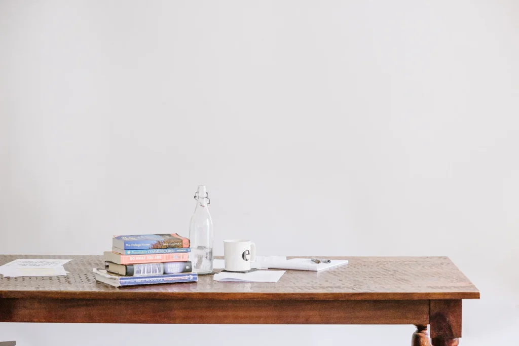 A wooden table with stacked books, a glass bottle, a white mug, a pen, and scattered papers, set against a plain white background.