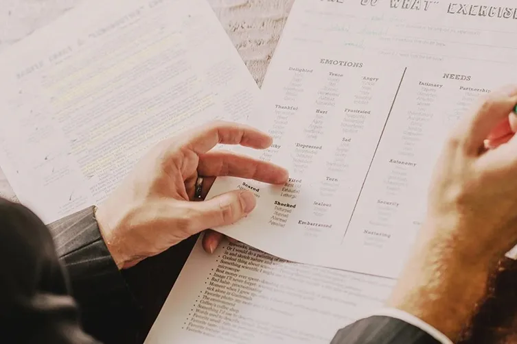Close-up of a person in a suit reviewing printed worksheets labeled "Emotions" and "Needs," highlighting and taking notes with a pen.