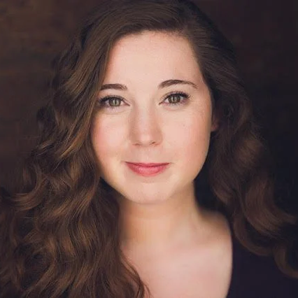 A woman with long, wavy brown hair and fair skin looks directly at the camera with a neutral expression against a dark background.
