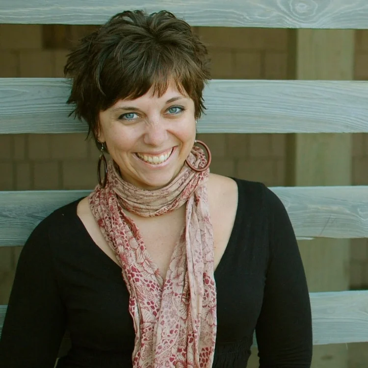Woman with short brown hair and blue eyes, wearing a black top, pink patterned scarf, and hoop earrings, standing in front of a wooden fence and smiling at the camera.
