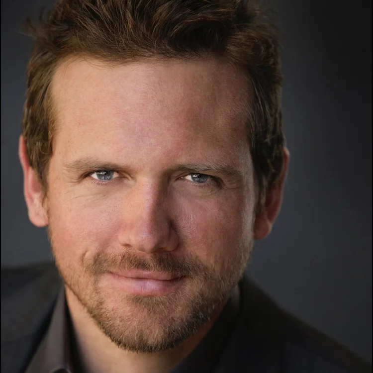 A man with short brown hair, light beard, and blue eyes looks directly at the camera, wearing a dark jacket against a dark background.