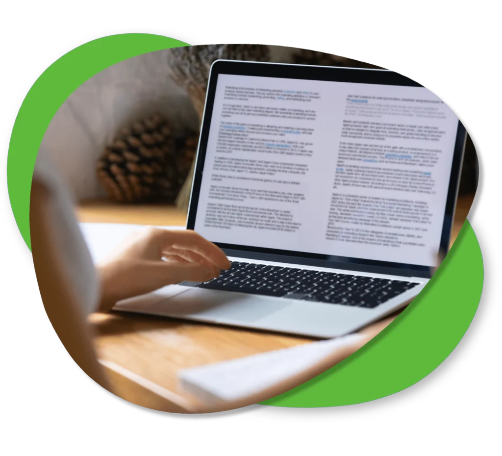 A person uses a laptop on a wooden desk, reading and typing text on the screen. Pinecones and other objects are visible in the blurred background.