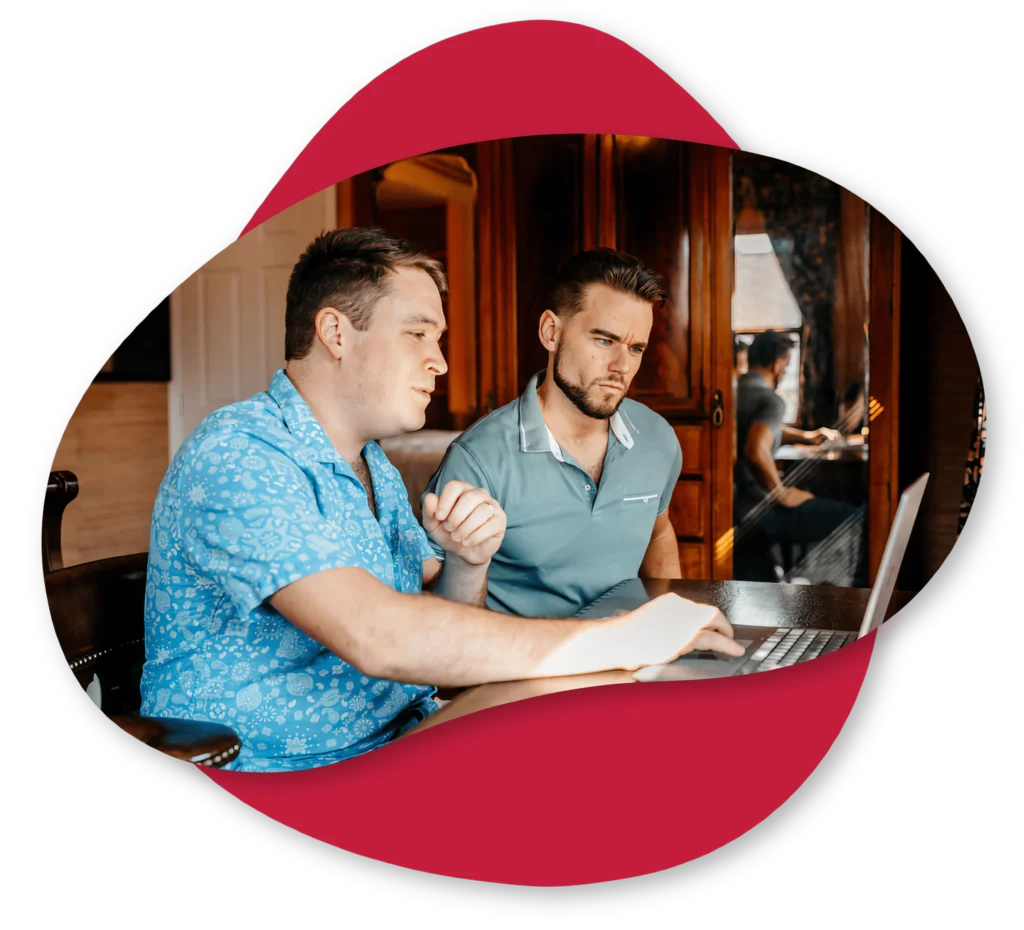 Two men sit at a table looking intently at a laptop screen, with one man pointing at the screen and the other watching closely.