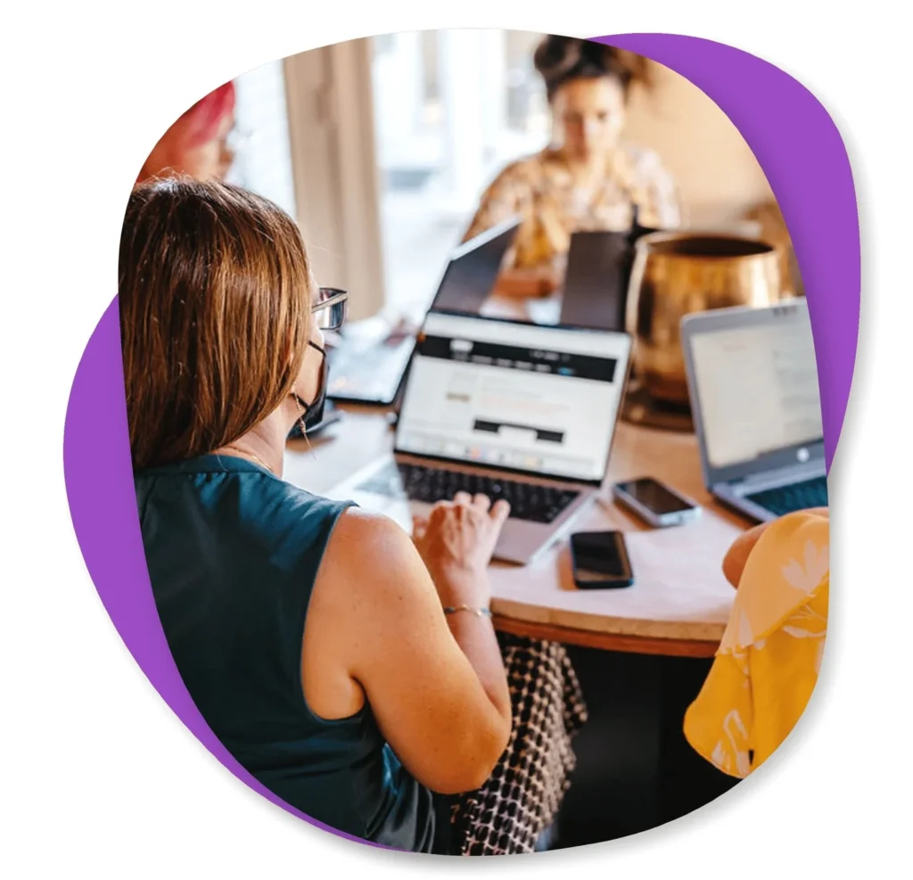 Several people sit around a table working on laptops, with smartphones and notebooks also on the table. The image is overlaid with a purple abstract shape.