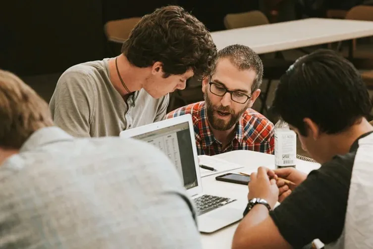 Four people sit around a table collaborating, with laptops and phones in use. Two of them are talking closely, while the others focus on their devices.