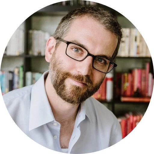 A bearded man wearing glasses and a light-colored shirt sits in front of a bookshelf filled with books.
