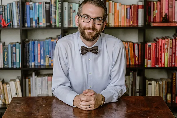 A man with a beard and glasses, wearing a light shirt and bow tie, sits at a wooden table in front of bookshelves filled with colorful books.