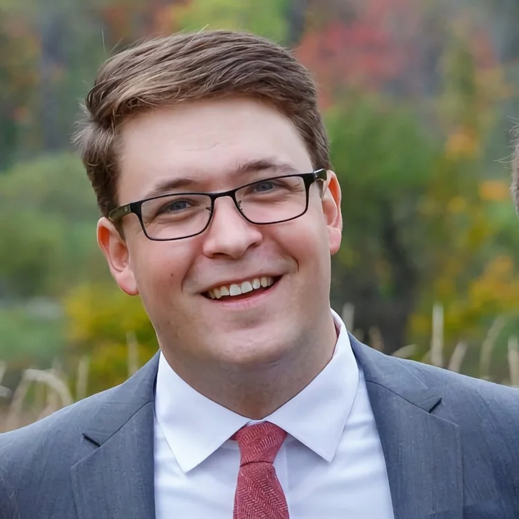 A man wearing glasses, a gray suit, white shirt, and pink tie smiles outdoors with blurred greenery and autumn foliage in the background.