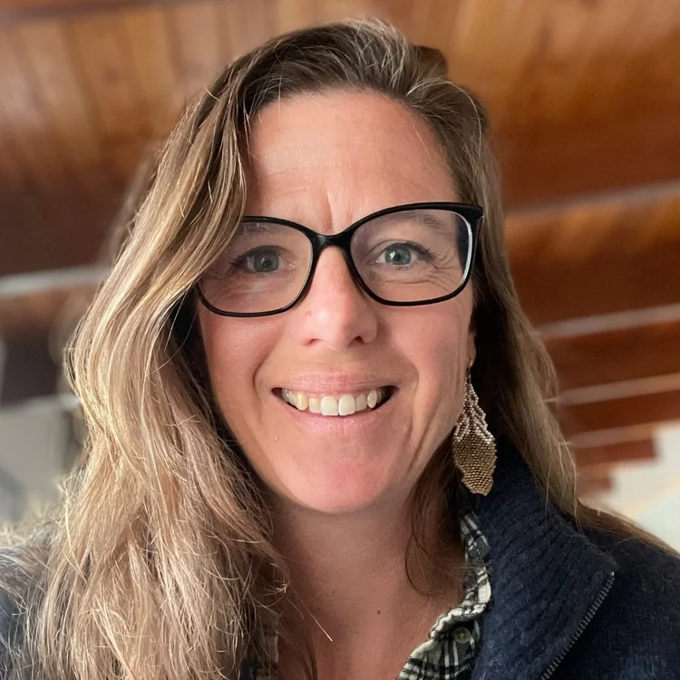A woman with long brown hair, wearing glasses, a patterned earring, and a dark sweater, smiles at the camera indoors with a wooden ceiling in the background.