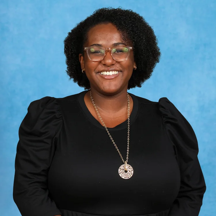 A woman with curly hair, glasses, and a black top smiles at the camera against a plain blue background. She is wearing a long necklace with a round pendant.