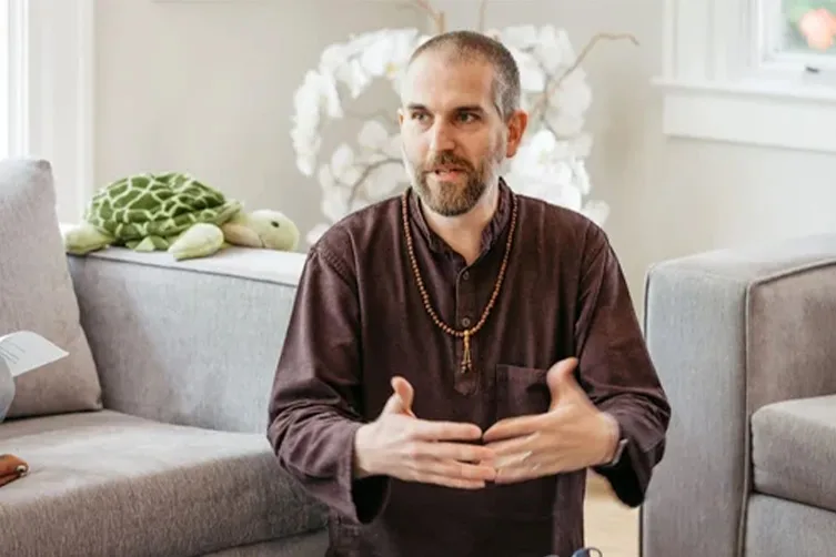 A man with a beard wearing a dark shirt and wooden bead necklace gestures with his hands while seated in a living room with a stuffed turtle on the couch behind him.