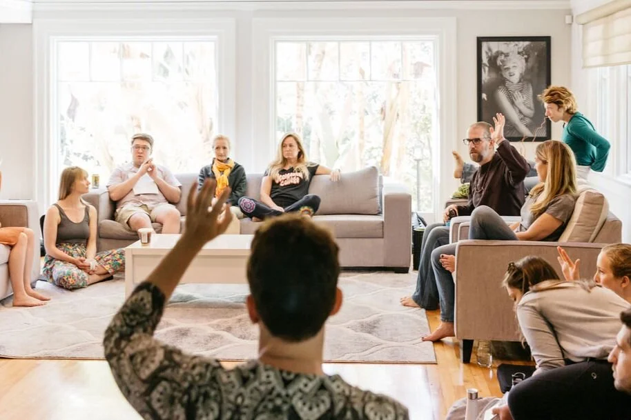 A group of people sit in a bright living room, some on sofas and others on the floor, with a few raising their hands.