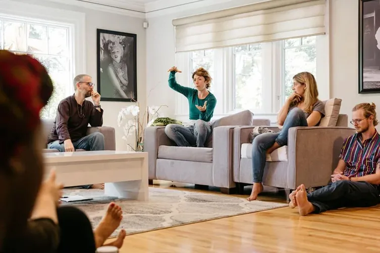 Four people sit and talk in a bright living room, with one person gesturing animatedly while others listen. A photo portrait hangs on the wall.