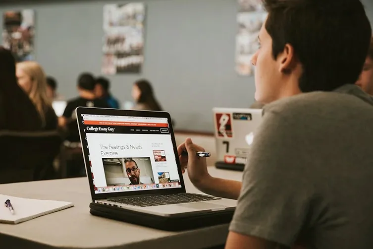 A student sits at a desk using a laptop, watching an educational video in a classroom with other students in the background.