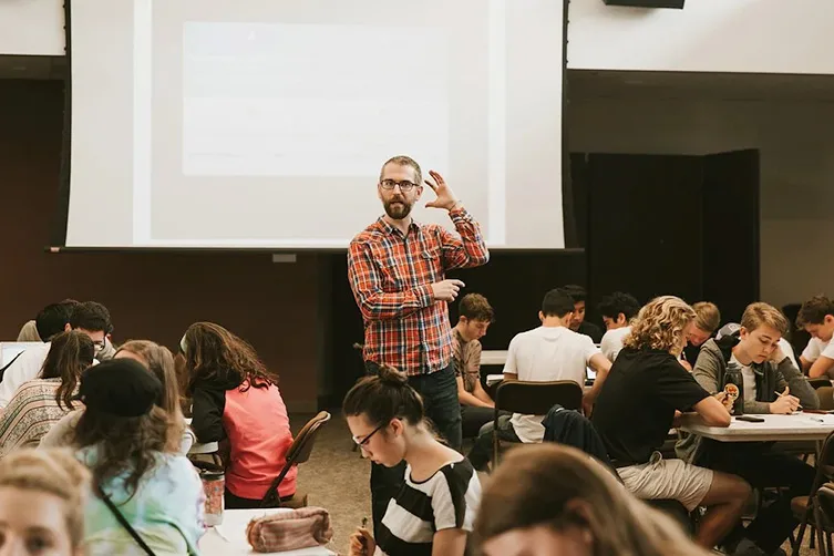 A man stands and gestures while speaking to students seated at tables in a classroom with a large projection screen behind him.