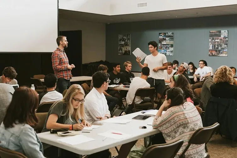 A classroom setting with students seated at tables, some writing and others listening to a standing student speaking while holding papers, and an instructor standing nearby.
