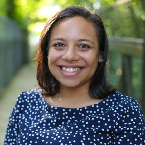 A woman with dark hair and a navy polka dot top smiles outdoors, with greenery and a blurred fence in the background.