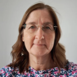 A middle-aged woman with brown hair, wearing glasses and a floral top, stands in front of a plain light background, looking at the camera with a neutral expression.
