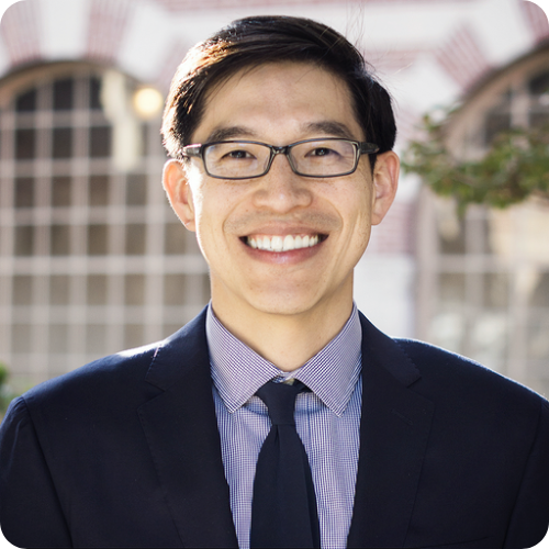 A man wearing glasses, a dark suit, and a tie smiles at the camera outdoors with a blurred building in the background.