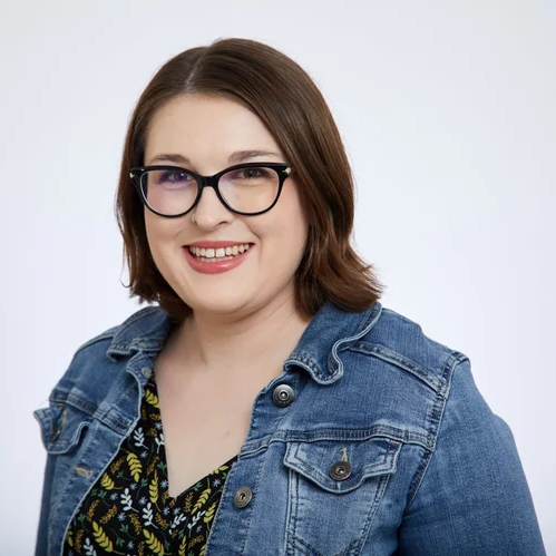 Woman with brown shoulder-length hair and glasses smiles at the camera, wearing a denim jacket over a patterned top against a plain white background.