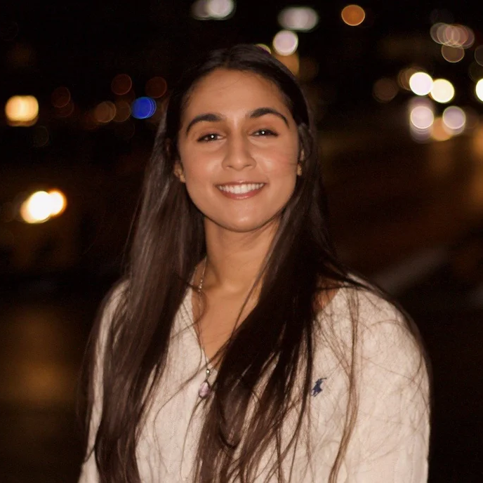 A woman with long dark hair wearing a light sweater stands outside at night, smiling at the camera with blurred city lights in the background.