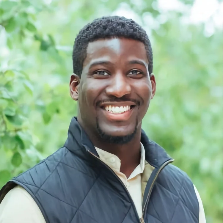 A man with short curly hair and a trimmed beard smiles at the camera, wearing a quilted dark vest over a light shirt, standing outdoors with green foliage in the background.