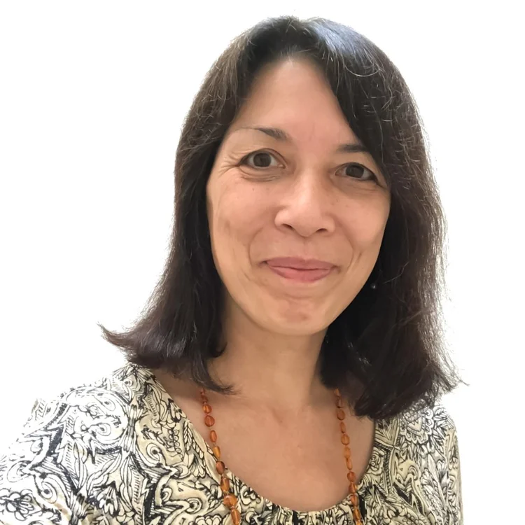 A woman with shoulder-length dark hair, wearing a patterned top and an amber beaded necklace, smiles at the camera against a plain white background.