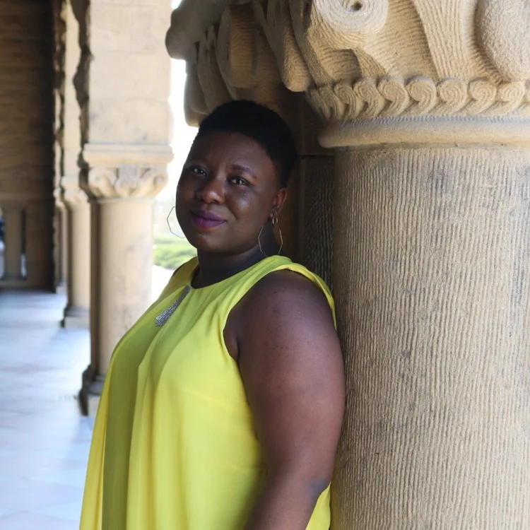 A person wearing a sleeveless yellow top stands indoors, leaning against a large stone column with intricate carvings.