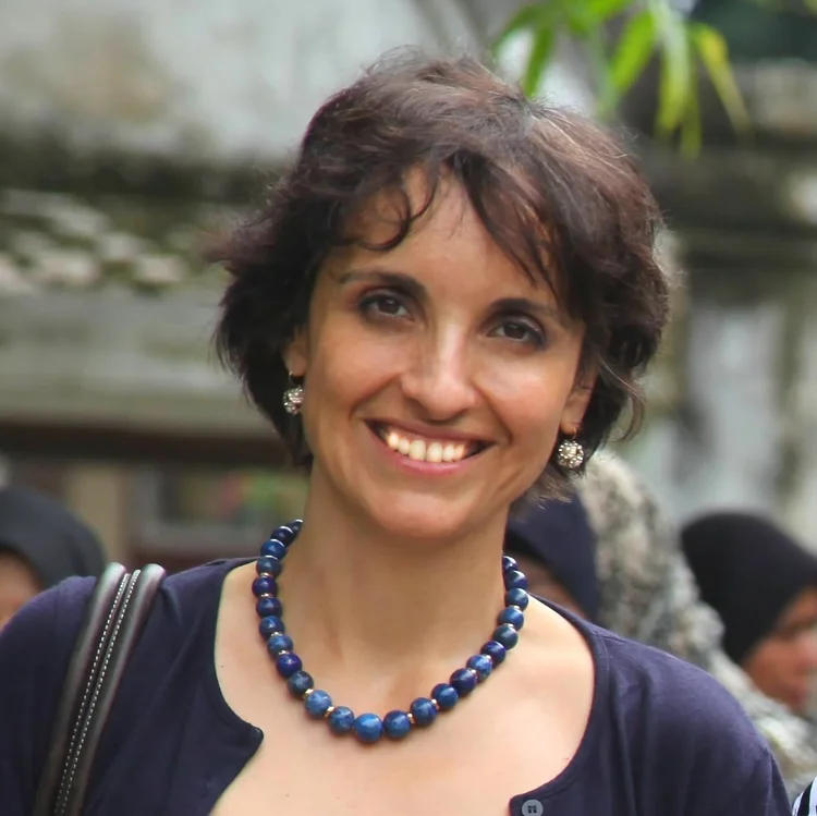 A woman with short dark hair, wearing a blue beaded necklace and earrings, smiles at the camera outdoors.
