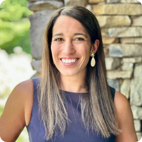 A woman with straight brown hair wearing a sleeveless navy dress and earrings stands in front of a stone wall, smiling at the camera.