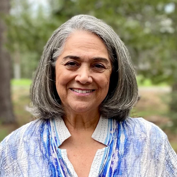 A woman with shoulder-length gray hair smiles outdoors, wearing a textured white and blue top. Green trees are visible in the background.