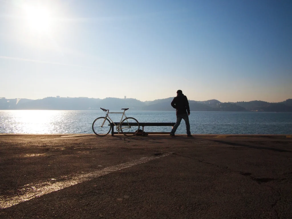 A person stands near a bicycle by the waterfront under a clear sky, with sunlight reflecting off the water and hills visible in the distance.