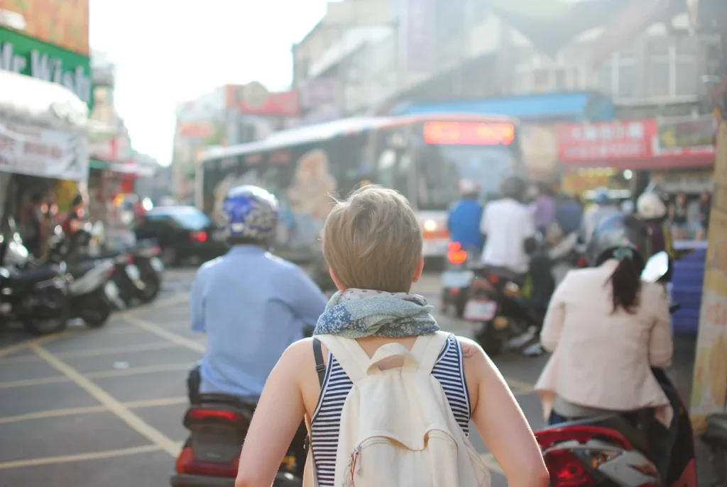 Person with a backpack stands among people on scooters in a busy urban street with traffic and buildings in the background.