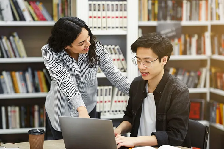 Two people in a library are working together at a table with a laptop; one is seated typing, and the other is standing, pointing at the screen and smiling. Bookshelves are in the background.