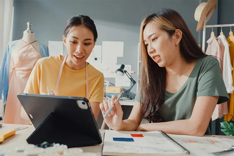 Two women sit at a desk with a tablet and sketchbook, discussing fashion designs in a workspace with clothing and sketches in the background.