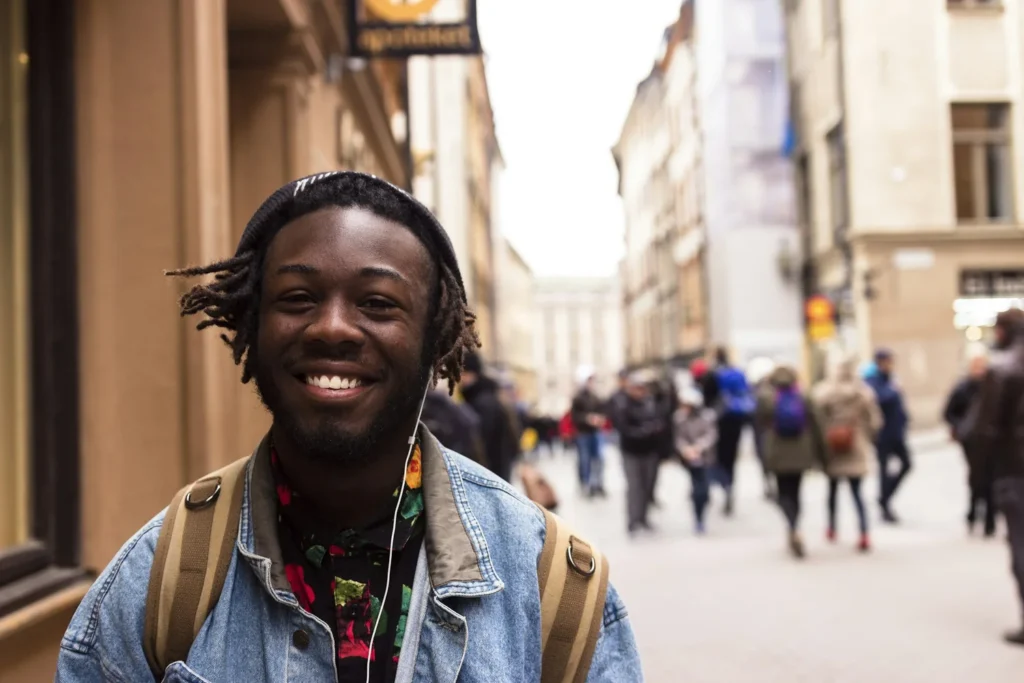 A young man with a backpack and headphones smiles at the camera on a busy city street with blurred pedestrians in the background.