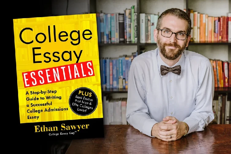 A man with glasses and a bow tie sits at a table with his hands folded. Next to him is the book "College Essay Essentials" by Ethan Sawyer. Bookshelves are in the background.