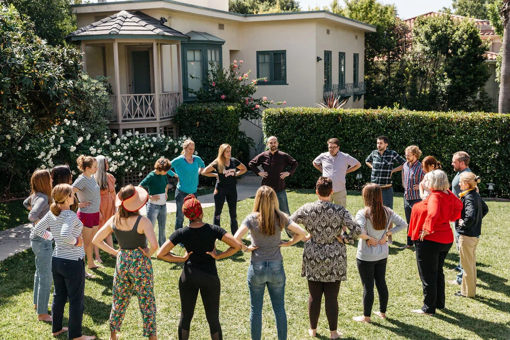 A group of people stand in a circle with hands on hips on a lawn outside a house, participating in an outdoor activity.