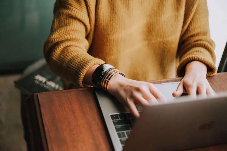 Person wearing a mustard sweater types on a laptop at a wooden desk, with a book partially visible beside them.