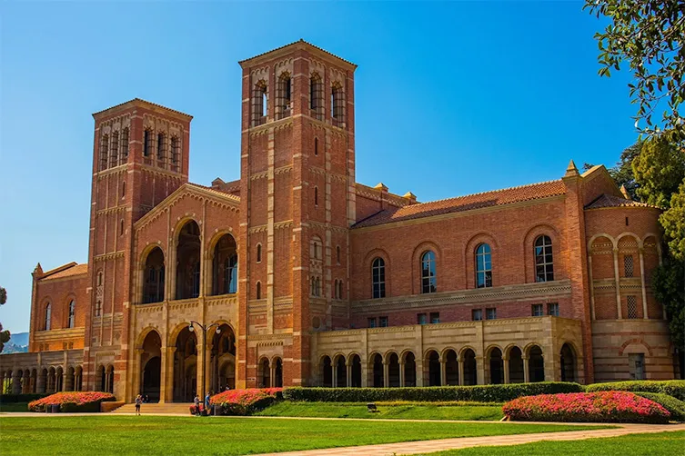 A large brick building with two towers, arched windows, and a landscaped lawn under a clear blue sky.