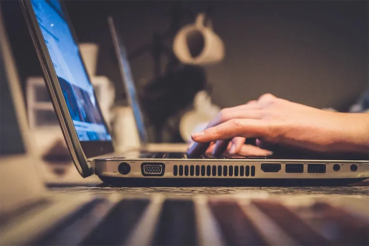A person types on a laptop keyboard, with another laptop and a blurred background visible.