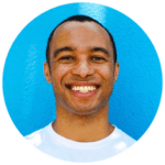 A person with short hair smiles at the camera, standing in front of a bright blue wall and wearing a white shirt.