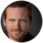A man with short brown hair and light facial hair looks directly at the camera, with a slight smile and a dark background.