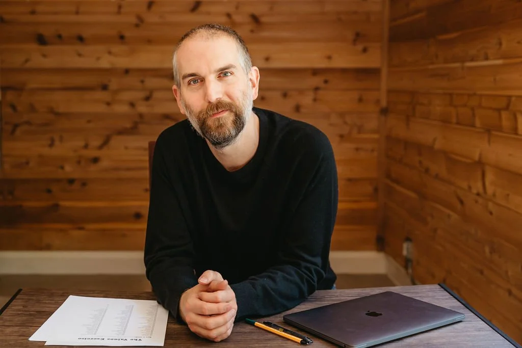 A man with a beard sits at a wooden desk with a laptop, pencil, and a sheet of paper with printed graphs, against a wood-paneled wall.
