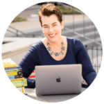 A woman with short brown hair sits outdoors at a table, smiling at the camera, with a laptop in front of her and a stack of books to her left.