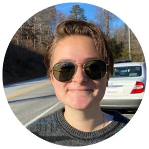 Person wearing sunglasses and a sweater, standing on a roadside in sunlight with trees and parked cars in the background.