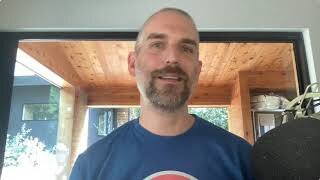 A man with a beard and short hair is smiling indoors near a window, wearing a blue T-shirt.