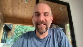 A man with a shaved head and beard, wearing a blue t-shirt, sits indoors in front of a window with wooden ceiling panels and greenery visible outside.