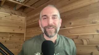 A man with a beard and mustache smiles at the camera while speaking into a microphone in a wooden-paneled room.